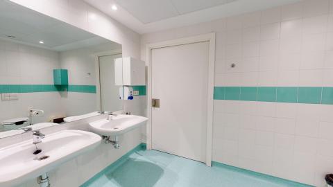 Baño. Cuarto de baño interior amplio y accesible, con paredes de azulejos blancos interrumpidas por una franja horizontal de baldosas de color verde agua. El suelo es de vinilo antideslizante en un tono azul claro uniforme. Cuenta con dos lavabos suspendidos sin pedestal y un gran espejo continuo empotrado en la pared que refleja la estancia. La luminosidad es artificial, blanca y homogénea, y destaca una puerta de madera lacada en blanco con manivela adaptada.
