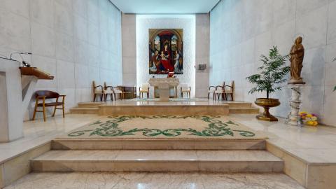 Capilla. Interior de una capilla solemne y minimalista, con paredes y suelo revestidos de mármol o piedra clara de tonos grisáceos y beige. Al fondo, sobre un altar elevado por tres escalones, destaca una gran pintura religiosa clásica enmarcada. A la derecha se aprecia una figura de la Virgen sobre una columna decorada y una planta verde en un macetero dorado. La estancia recibe una luminosidad clara y difusa que resalta una alfombra central con motivos ornamentales verdes.