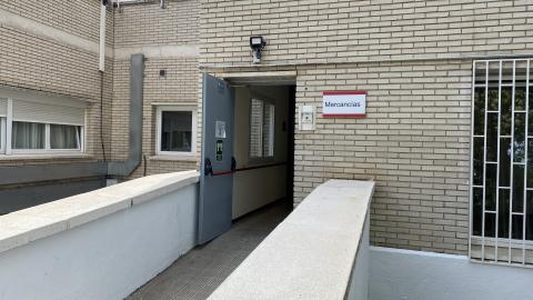 Acceso exterior a una zona secundaria identificada con un cartel rojo y blanco donde se lee "Mercancías". El entorno muestra una rampa de suelo técnico gris flanqueada por muretes blancos de hormigón. La fachada del edificio es de ladrillo visto ocre claro con una ventana enrejada a la derecha. La luminosidad es de un día nublado, proporcionando una luz diurna suave y sin sombras marcadas. Destaca una puerta metálica gris entreabierta hacia el pasillo interior.