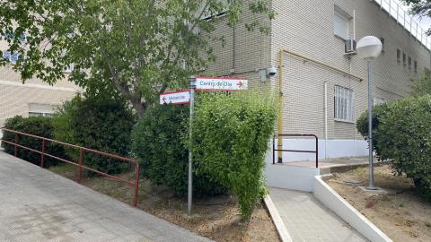 Entorno ajardinado exterior con rampas de hormigón para favorecer la accesibilidad peatonal, protegidas por barandillas metálicas de color granate. En el centro destaca un poste con flechas de señalización blancas y rojas que apuntan hacia "Centro de Día" y "Mercancías". El entorno está rodeado de frondosos árboles y arbustos verdes bajo una luz natural diurna y clara. Al fondo se aprecia la fachada de ladrillo del edificio principal con una farola de bola blanca.