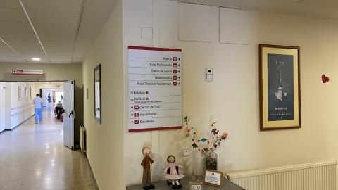 Una esquina de un pasillo interior con paredes de color beige y suelo de terrazo claro pulido. La iluminación es suave y proviene del techo. En la pared central destaca un gran panel blanco de directorio con texto y flechas rojas. Debajo del panel, sobre un mueble recibidor gris con detalles rojos, destacan dos muñecos de trapo artesanales junto a un adorno con forma de árbol de hojas coloridas.