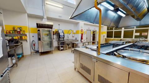 Cocina. Cocina industrial amplia con equipamiento de acero inoxidable y azulejos blancos con una franja amarilla. Destaca una gran isla central con fuegos y campana extractora iluminada. Al fondo se aprecian hornos profesionales y estanterías con utensilios bajo una luz blanca y clara.