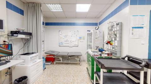 Consulta de enfermería. Consulta médica o de enfermería muy iluminada, con paredes de azulejo blanco decoradas con una franja azul. Destaca una camilla de exploración gris en el centro. La sala incluye un carro de curas verde, un escritorio con ordenador y un panel de corcho con notas al fondo.