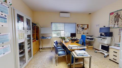 Consulta médica. Despacho médico con luz natural suave proveniente de una ventana central con persianas. Las paredes son de color crema y el suelo de terrazo claro. Destaca una mesa de escritorio de madera con sillas azules para pacientes, armarios de almacenamiento y un póster de anatomía humana en la pared derecha.