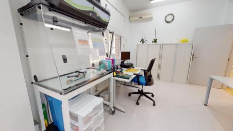Depósito de medicamentos. Depósito de medicamentos con paredes blancas y suelo claro, muy bien iluminada. Destaca en primer plano una cabina de flujo laminar o vitrina de seguridad de policarbonato transparente. Al fondo se observa un escritorio con ordenador, silla azul y armarios archivadores blancos.