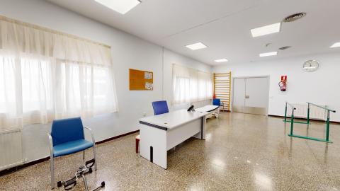 Fisioterapia. Sala de fisioterapia amplia y luminosa con paredes blancas y suelo de terrazo brillante. Destacan unas barras paralelas de marcha a la derecha y una camilla al fondo. En el centro hay una mesa de despacho blanca con sillas azules y una espaldera de madera sujeta a la pared.