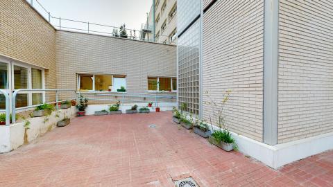 Patio. Un espacio al aire libre con mesas y sillas para actividades. Paredes blancas y muy soleado. Cuenta con una zona con jardineras y plantas. Diseñado para fomentar la autonomía y la creatividad.