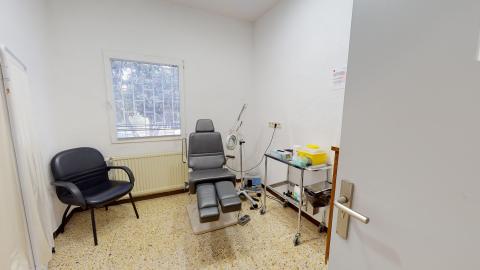 Consulta de podología. Consulta de podología de paredes blancas iluminada por una ventana central con rejas y vistas a árboles. En el centro destaca un sillón médico articulado de color gris oscuro, acompañado a la izquierda por una silla de confidente negra y a la derecha por una mesa auxiliar de acero inoxidable con material sanitario. El suelo es de terrazo amarillento.