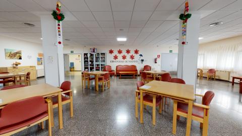 Sala de actividades. Amplia sala común de actividades con mesas y sillas de madera con tapicería granate distribuidas por todo el espacio. Al fondo hay un área de descanso con sofás tapizados en color teja y un mueble de biblioteca con libros. La estancia cuenta con pilares blancos decorados con adornos colgantes, un suelo de terrazo oscuro y ventanas con cortinas claras a la derecha que aportan luminosidad.