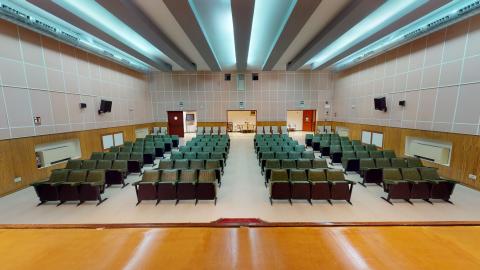 Salón de actos. Perspectiva elevada del salón de actos tomada desde el escenario de madera hacia la zona del público. Se aprecian las filas de butacas de color verde oscuro distribuidas simétricamente en una sala de paredes beige con zócalo de madera. Al fondo se ven las puertas de salida y el techo técnico gris con sistemas de iluminación y climatización longitudinales.