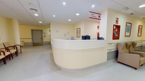 Enfermería. Zona de paso interior y control de enfermería con iluminación artificial empotrada en el techo técnico blanco. La estancia tiene paredes de color crema, suelo vinílico gris claro y pasamanos integrados. Destaca el mostrador central semicircular de color blanco y crema, coronado por un cartel colgante que indica "Control Enfermería", flanqueado por sillones de descanso de color beige a ambos lados.