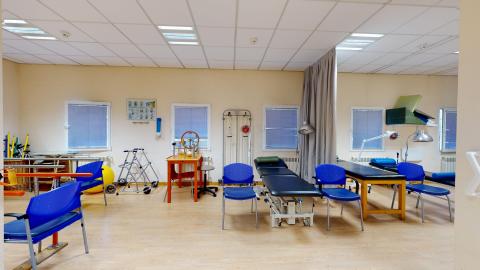 Fisioterapia. Sala de rehabilitación muy iluminada mediante fluorescentes en el techo y varias ventanas con persianas. Las paredes son de color beige claro y el suelo imita madera clara. Destaca la variedad de equipamiento terapéutico: varias camillas de exploración oscuras, sillas de un llamativo color azul eléctrico, un andador, espalderas y una pelota de pilates amarilla al fondo.
