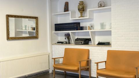 Sala de espera vestíbulo principal. Una acogedora zona de espera con dos sillones de cuero naranja y madera frente a una pared de ladrillo blanco. Encima hay estanterías flotantes con libros, jarrones y máquinas de escribir antiguas. Un gran espejo con marco dorado cuelga a la izquierda sobre un radiador. El suelo es de terrazo oscuro.