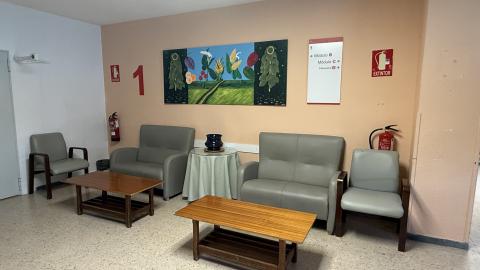 Sala de espera visitas planta habitaciones. Un área de descanso con dos sofás de cuero gris y dos sillones a juego organizados alrededor de dos mesas de centro de madera. La pared trasera es de color melocotón y tiene un gran cuadro colorido de un paisaje. Un extintor rojo está colgado en la pared cerca del cuadro. El suelo es de terrazo claro.