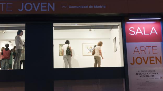 Vista desde el exterior de una estancia de la sala de arte joven iluminada, con visitantes conociendo una exposición