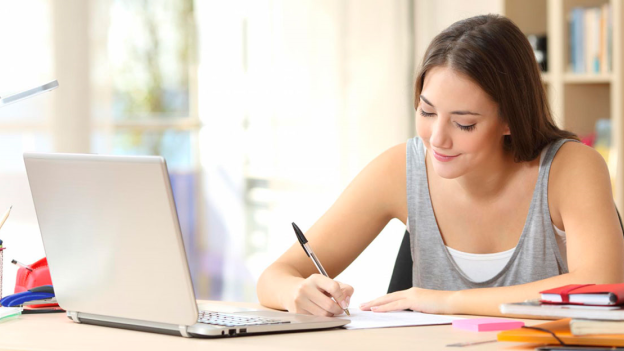 Chica joven escribiendo en un cuaderno, con un portátil al lado