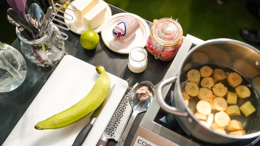 Plátano verde sobre tabla de cortar junto a una olla con rodajas de plátano cocinándose en una placa de inducción