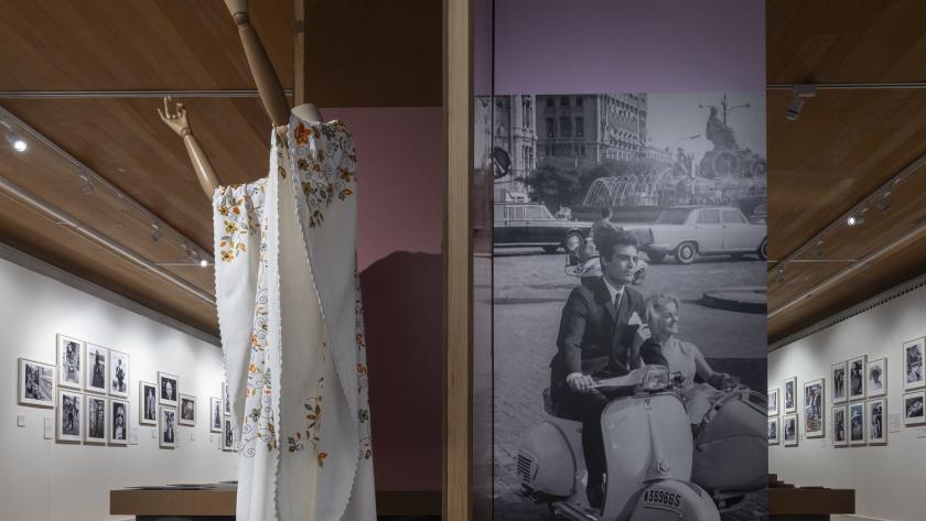 Vista de la sala expositiva, en primer plano una pieza de indumentaria femenina y una fotografía en blanco y negro de dos personas en sidecar
