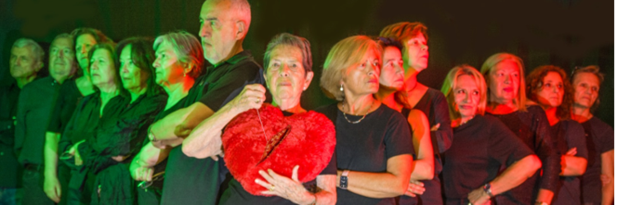Varias personas vestidas de negro y en forma de uve miran hacia los lados. La persona del centro mira hacia el frente y sostiene un corazón roto de peluche