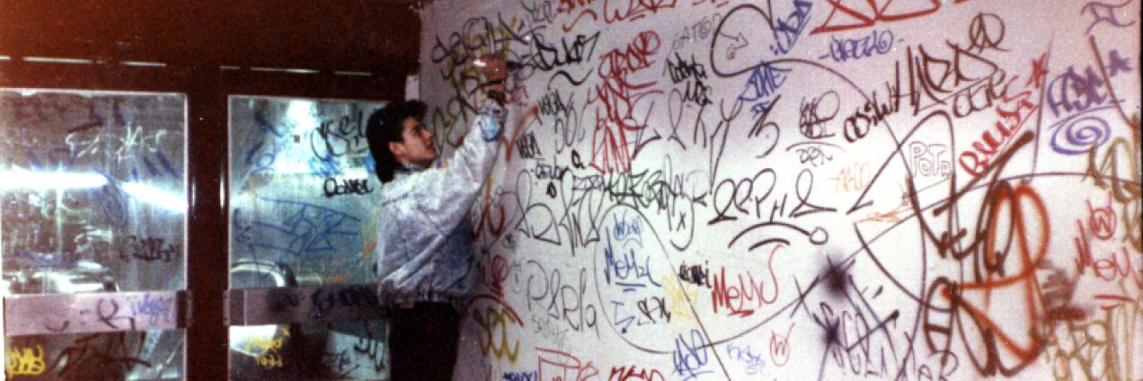 Fotografía de un chico vestido con moda de los 90 firmando una pared llena de grafitis