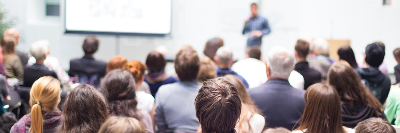 Un grupo de personas sentadas en un aula, escuchando a un conferenciante que está al fondo, delante de una pared y de una pizarra