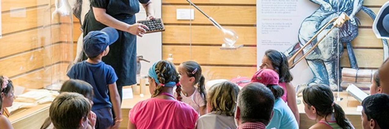 Niños y niñas en el centro de interpretación mirando a la educadora de la actividad que les explica cómo se hace el vídrio