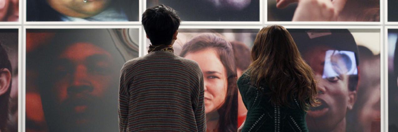 Dos mujeres sentadas de espaldas al espectador y mirando una pared llena de fotografías con retratos en color de personas anónimas