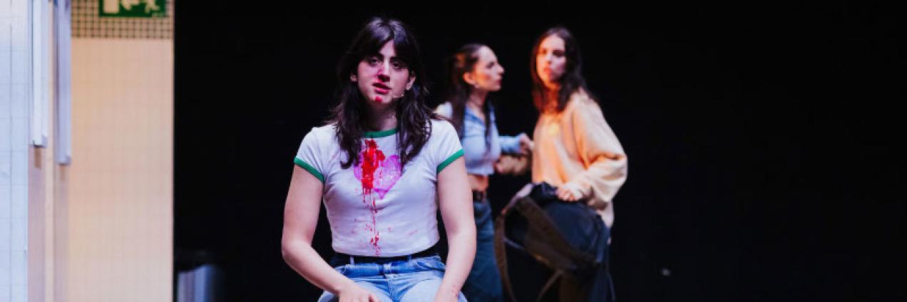 imagen de tres chicas en escena, una de ella está sentada y sangra por la nariz, tras ella las otras dos hablan 
