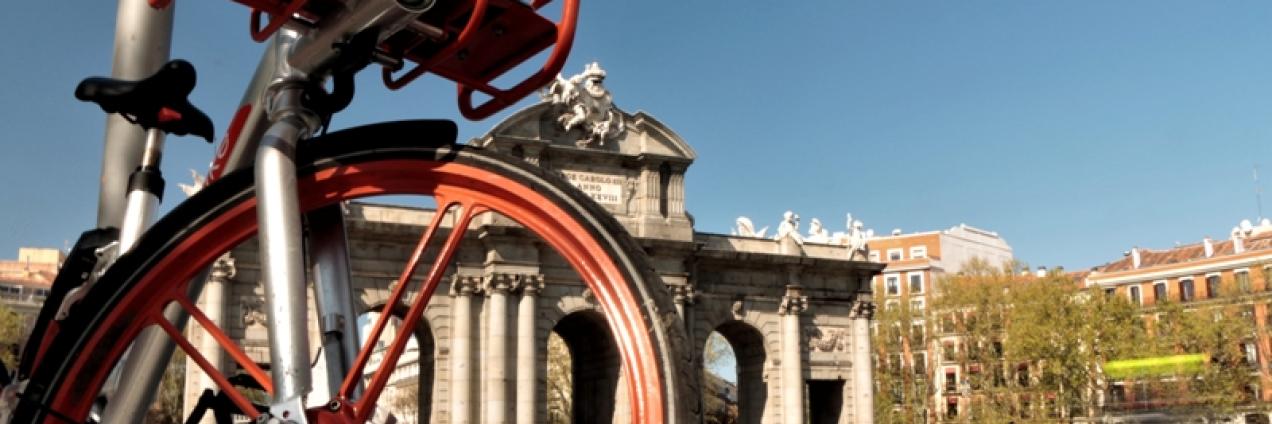 IMAGEN DE LA PUESTA DE ALCALÁ VISTA TRAS LOS RADIO DE LA RUEDA DE UNA BICICLETA ROJA