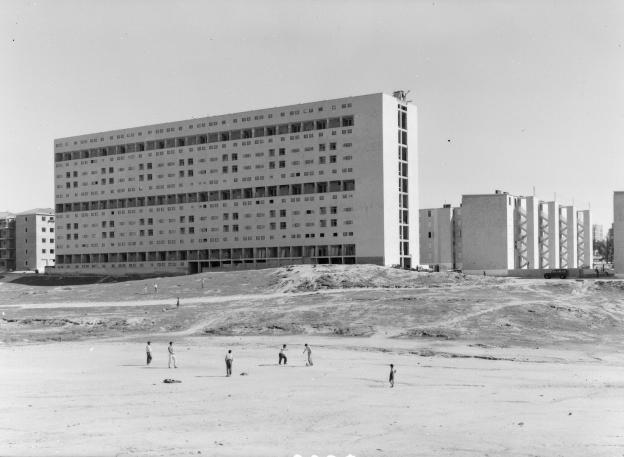 Imagen de un descampado con niños jugando al fútbol y edificios al fondo