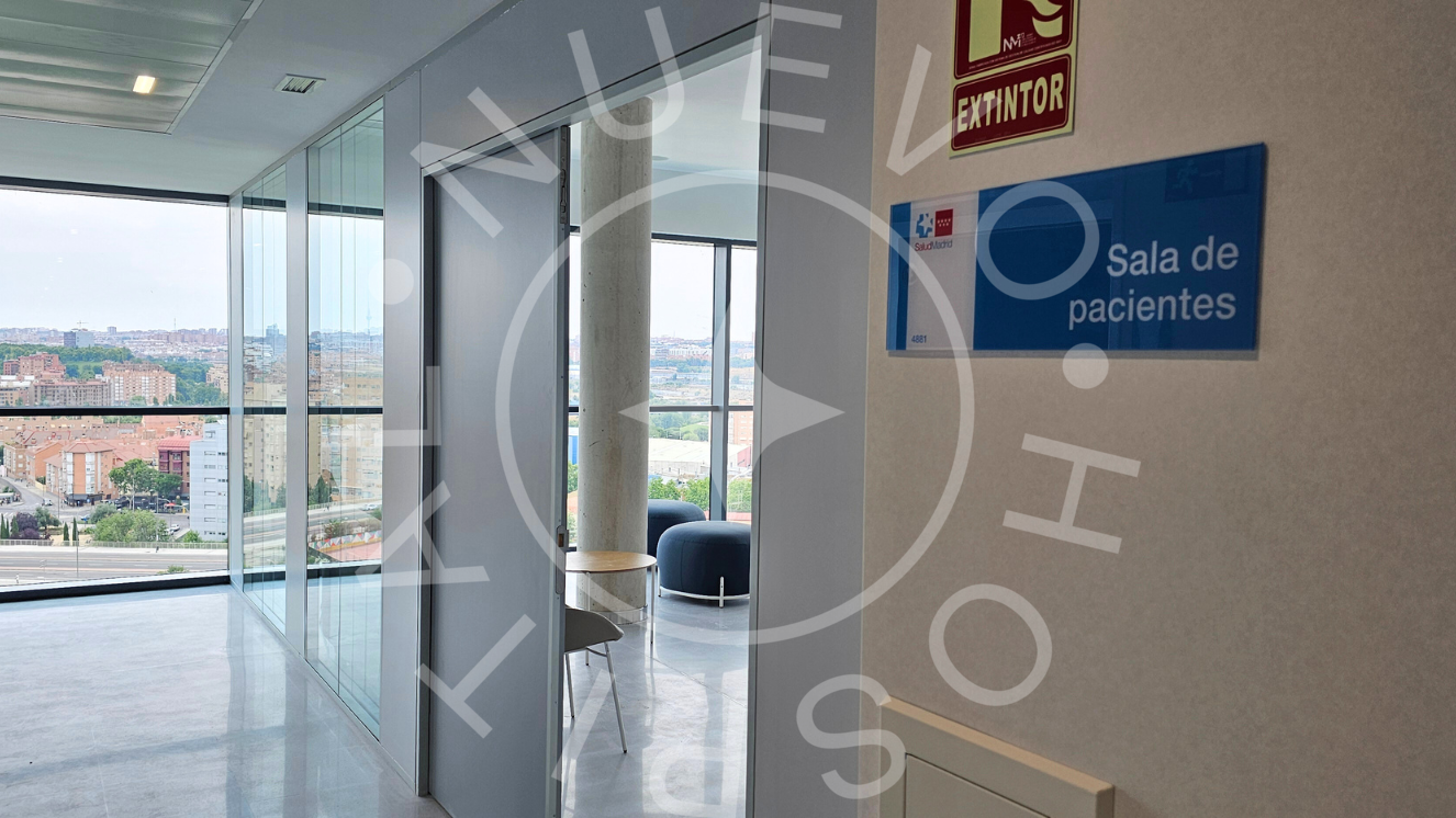 Acceso a sala de espera hospitalización adultos con cartel en pared que indica sala de espera y al fondo a la derecha puerta abierta por la que se entreveen unos sofás y al fondo del pasillo ventanal con paisaje 