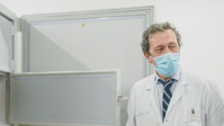 un profesional con bata y un hombre con traje gris, ambos de medio cuerpo delante de una gran nevera de laboratorio abierta