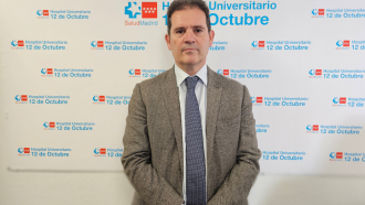 Hombre con traje chaqueta gris y corbata azul posando con fondo de panel con logos