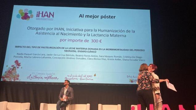 Plano general de dos mujeres que se abrazan en un escenario, una de ellas sostiene un diploma en una mano