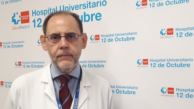 Retrato de hombre con barba blanca recortada y gafas con camisa, corbata y bata blanca sobre fondo de logos