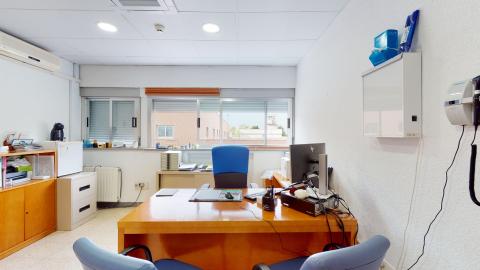 Consulta médica. Consulta en color blanco. Al fondo hay ventanas de izquierda a derecha. Debajo de éstas, una consola. En el muro izquierdo una archivero de metal y una consola de madera. Al centro el escritorio de madera con sillas azules. En el muro derecho una lámpara para ver radiografías.
