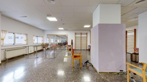 Fisioterapia Amplio espacio en blanco y morado claro. En el muro izquierdo hay dos amplias ventanas. Al fondo un gran espejo de cuerpo completo. Al fondo derecha y muro derecho barras de madera. En el centro apoyos para andar y estaciones para ejercitar las piernas.