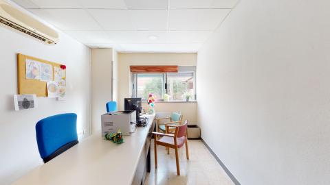 Consulta de psicología. Sala alargada en blanco. Al fondo una amplia ventana que da el exterior, se alcanza a ver vegetación. A lo largo está el escritorio con sillas a ambos lados.