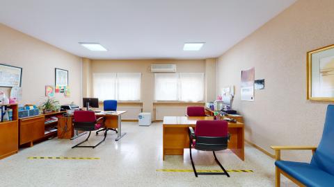 Trabajo social. Sala rectangular en tonos marrón claro y blanco. Al fondo dos amplias ventanas con cortinas blancas traslúcidas. En el muro de la izquierda muebles bajos de madera. Alineados con las ventanas, hay dos escritorios de atención de madera.