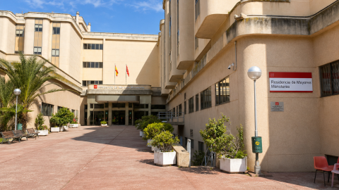 Vista del acceso a la residencia. Es una construcción de varios edificios en beige con un amplio pasillo al centro con asientos en el exterior y una palmera.
