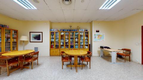 La sala cuenta con paredes de tono amarillo claro, suelo de terrazo grisáceo y mobiliario de madera que incluye varias estanterías repletas de libros, dos mesas de estudio de madera clara con sillas de cuero marrón, un cuadro en la pared y un mapa de España colgado junto a una mesa auxiliar de oficina.