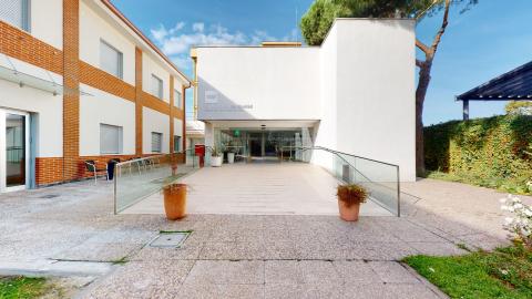 Jardín. Entrada principal moderna y muy luminosa bajo un cielo despejado. Destaca un bloque central blanco con letras plateadas de la "Comunidad de Madrid" y una amplia rampa de baldosas claras flanqueada por barandillas de cristal transparente. A la izquierda, un edificio de dos plantas combina fachada blanca con franjas de ladrillo visto arcilloso. Dos maceteros de arcilla con plantas bordean el camino y a la derecha se aprecia un jardín con un gran pino.