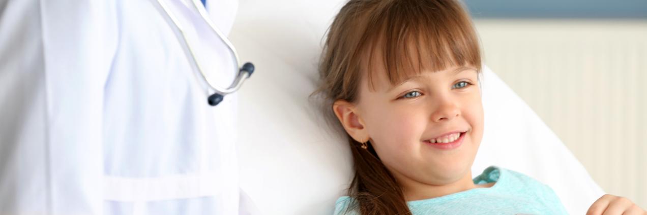 Paciente infantil recostada en una cama hospitalaria, con una persona sanitaria al lado vestida con bata y fonendoscopio, en un entorno clínico.