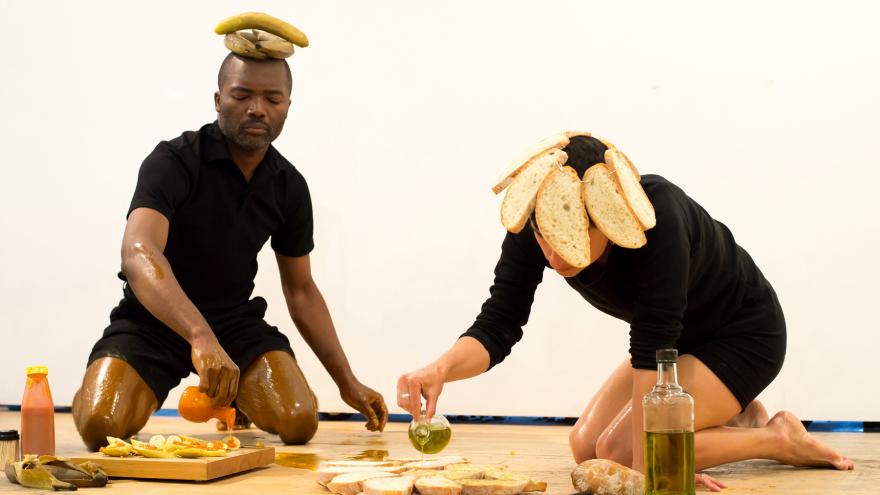 Un hombre con plátanos en la cabeza y una mujer con rebanadas de pan, preparan plátanos y pan para comer