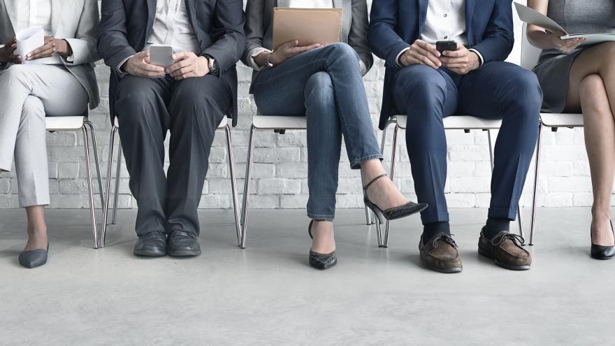 Five people are sitting waiting to be attended to in a personnel selection process. The two men are reading their cell phones, a woman is holding a