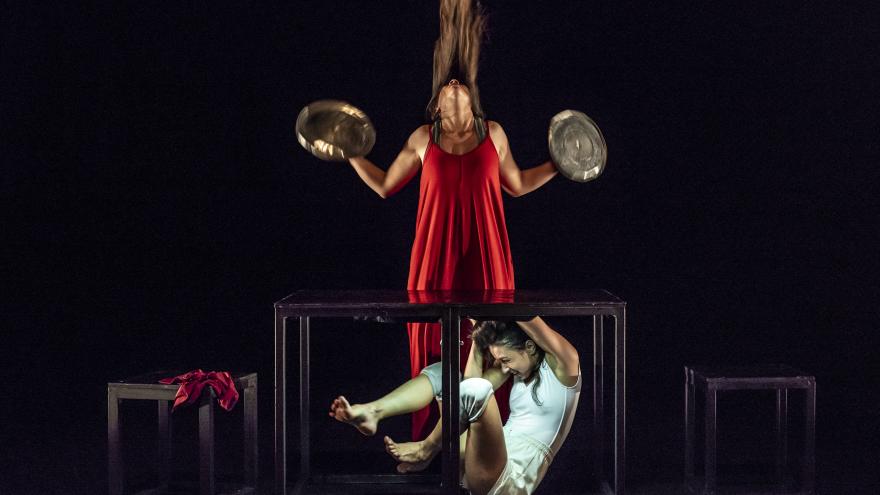 Dos mujeres hacen una performance. Una de ellas está encerrada en una caja de cristal y la otra, vestida de rojo, toca unos platillos de metal.