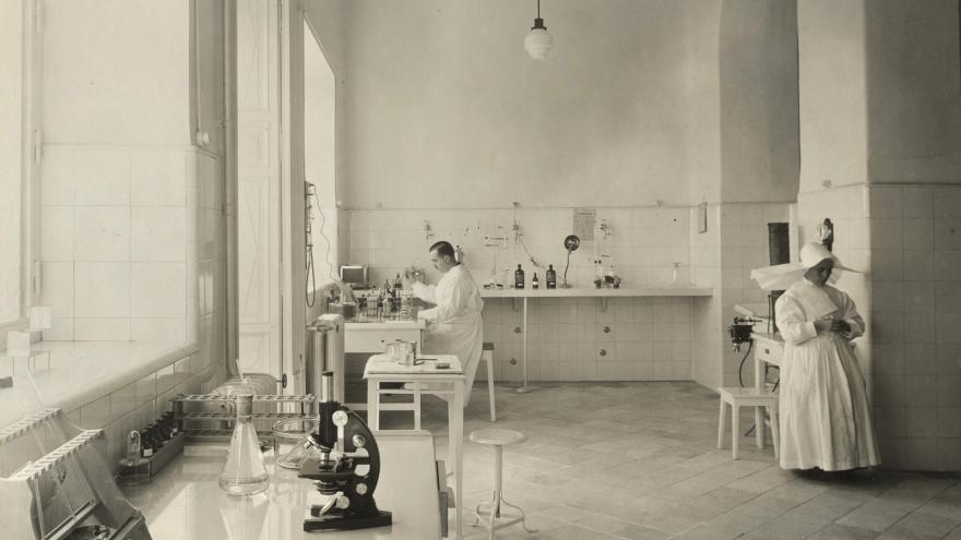 laboratorio médico con mesa y microscopio en segundo plano, médico al fondo de la estancia y enfermera a la derecha de la fotografía