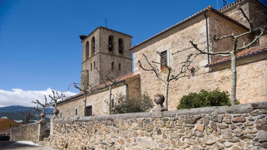 Fachada exterior de una iglesia con torre campanario rectangular rodeada por un muro de piedra