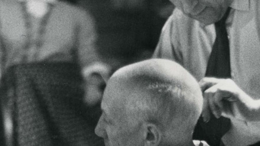 Black and white photograph of David D. Duncan in which Picasso appears seated while his barber Eugenio Arias cuts his hair