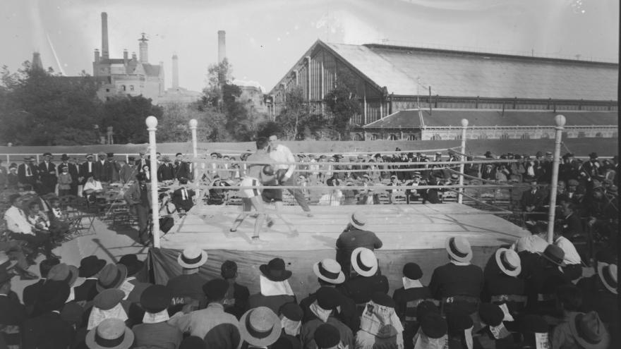 Foto en blanco y negro con un ring de boxeo y público observando el espectáculo